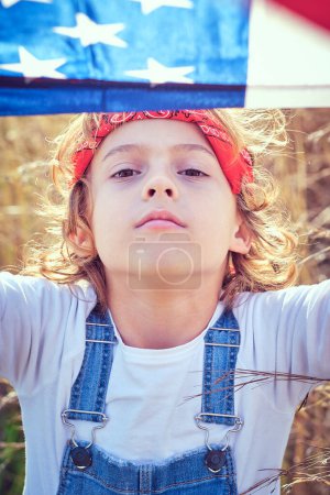 Foto de Adorable niño preadolescente cubriendo la cabeza con la bandera de Estados Unidos de América mientras descansa en el campo cubierto de hierba en el día soleado y mirando a la cámara - Imagen libre de derechos
