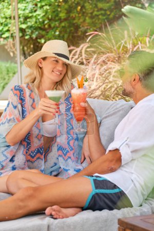 Téléchargez les photos : Vue latérale de l'homme adulte joyeux assis avec la jambe pliée et tenant verre à cocktail vers une femme souriante avec boisson aux fruits tout en se regardant - en image libre de droit