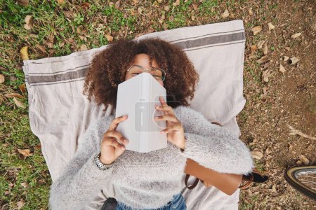 Téléchargez les photos : D'en haut de la femme afro-américaine en vêtements décontractés reposant sur une couverture sur l'herbe et couvrant le visage avec livre dans un parc vert le week-end - en image libre de droit