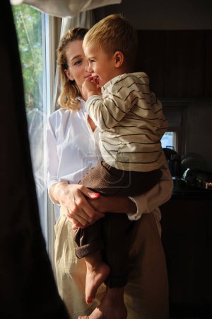 Foto de Joven madre en blusa blanca de pie cerca de la ventana y mirando suavemente y abrazando al hijo con suéter a rayas tomando un aperitivo en la habitación oscura - Imagen libre de derechos