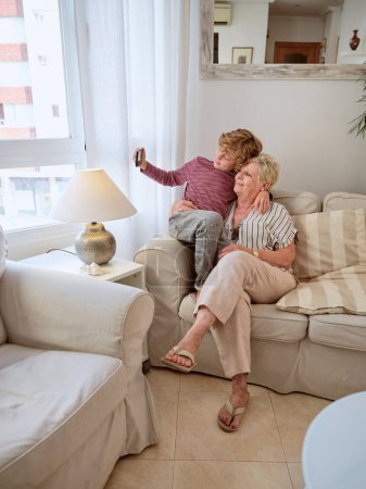 Téléchargez les photos : Vue latérale complète du corps de la grand-mère et du petit enfant assis sur le canapé et s'amusant ensemble dans un salon confortable à la maison - en image libre de droit