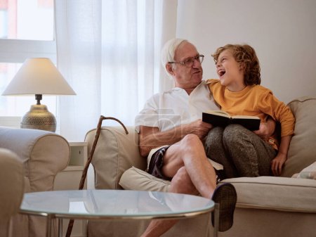Téléchargez les photos : Garçon joyeux avec grand-père lecture livre intéressant tout en passant du temps ensemble sur un canapé confortable dans le salon de la lumière à la maison - en image libre de droit