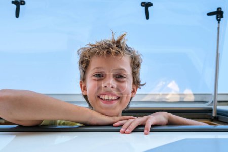Téléchargez les photos : D'en bas garçon optimiste souriant joyeusement et regardant la caméra tout en regardant par la fenêtre de la caravane contre le ciel bleu pendant le voyage sur la route - en image libre de droit