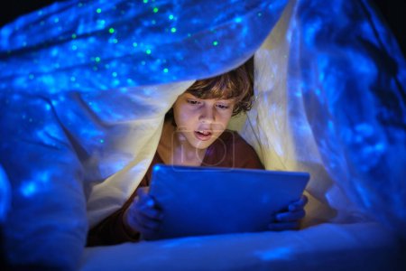 Téléchargez les photos : Garçon concentré naviguant tablette moderne tout en étant assis sous la couverture avec des lumières lumineuses dans la chambre sombre la nuit à la maison - en image libre de droit