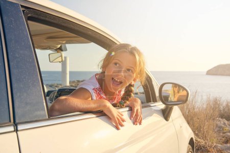 Foto de Chica rubia sacando la cabeza por la ventana de un coche estacionado frente al mar con una expresión feliz y el reflejo de su madre en el espejo lateral del coche - Imagen libre de derechos