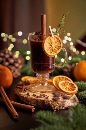 Foto de Copa de vino caliente alcohólico servido con palo de canela y rebanada de naranja seca colocada sobre fondo borroso en sala de luz - Imagen libre de derechos