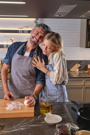 Wife hugging positive husband cutting onion on cutting board and looking at camera while cooking together in modern light kitchen