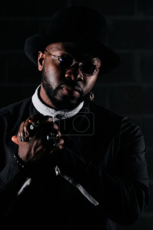Photo for Serious African American male model in black outfit with sunglasses looking away while standing against brick wall in dark room - Royalty Free Image