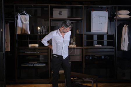 Foto de Hombre adulto en camisa blanca de pie delante del armario y poniéndose pantalones negros en luz tenue - Imagen libre de derechos