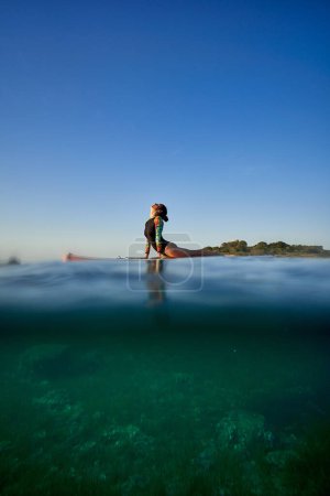 Photo for Side view full body of anonymous woman in swimwear doing Bhujangasana while practicing yoga on paddleboard in sea with clear turquoise water under cloudless evening sky - Royalty Free Image