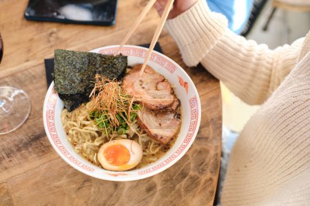 Photo for From above crop unrecognizable person eating appetizing ramen with wooden chopsticks served on wooden table - Royalty Free Image