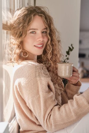 Téléchargez les photos : Vue latérale de la jeune femme aux cheveux blonds bouclés regardant la caméra tout en étant assis près de la fenêtre avec une tasse de thé - en image libre de droit