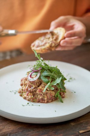 Photo for Unrecognizable person sitting at wooden table and spreading delicious steak tartare served with rucola on crouton at home - Royalty Free Image