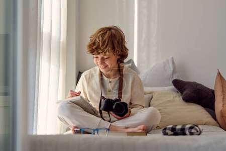 Foto de Escolar sonriente con cámara fotográfica profesional tomando notas en el cuaderno mientras está sentado con las piernas cruzadas en la cama en casa - Imagen libre de derechos