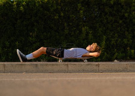 Foto de Vista lateral a nivel del suelo del niño preadolescente acostado en el monopatín en la calle y disfrutando de la puesta de sol en verano - Imagen libre de derechos