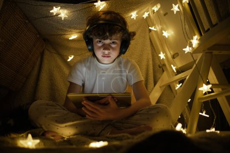 Photo for Low angle full body of boy in pajama and wireless headphones watching video on tablet sitting in playing tent decorated with glowing garland - Royalty Free Image
