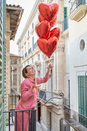 Photo for Side view of cheerful lady in sleepwear looking up at bunch of heart shaped balloons while standing on hotel balcony - Royalty Free Image