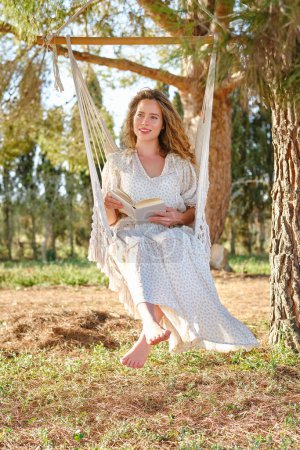 Photo for Full body positive woman smiling and looking away while relaxing on hammock on branch of coniferous tree and enjoying book on sunny day - Royalty Free Image