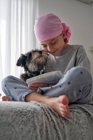 Photo for Low angle of Miniature Schnauzer dog sniffing face of smiling boy in pink bandana of breast cancer awareness campaign writing in notepad on sofa - Royalty Free Image