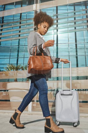 Photo for Side view of young African American female in trendy outfit and boots with bag walking to airport and looking down - Royalty Free Image