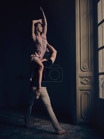 Photo for Full body of ballerina sitting on shoulder of faceless male dancer while dancing ballet dance in room with dim light - Royalty Free Image
