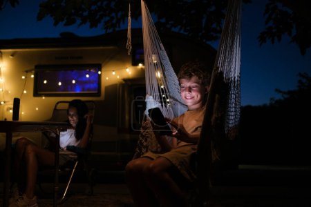 Foto de Niño contento que navega el teléfono móvil mientras que descansa en la hamaca cerca de RV y de la madre en la noche oscura en campo - Imagen libre de derechos