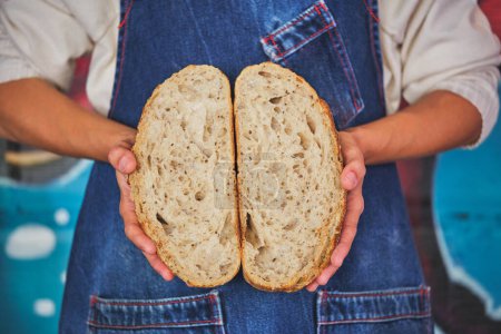 Téléchargez les photos : Détail photo des mains féminines tenant un pain artisanal rompu en deux avec un tablier de cuisine bleu. - en image libre de droit
