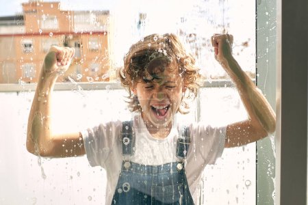 Téléchargez les photos : Enfant excité avec les yeux fermés s'amuser et crier tout en faisant la routine du ménage et laver les fenêtres et le balcon à la maison - en image libre de droit