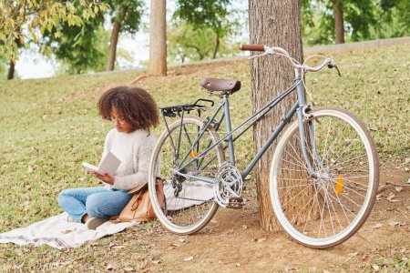 Photo for Full body of concentrated African American woman in casual clothes sitting sitting on blanket near bicycle and reading interesting story in green park - Royalty Free Image