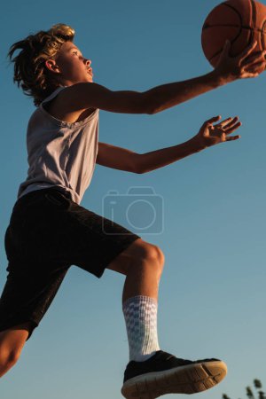 Téléchargez les photos : De dessous vue latérale du garçon énergique lançant la balle tout en jouant au basket-ball contre le ciel bleu - en image libre de droit