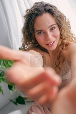 Photo for Cheerful young female with curly hair smiling and holding out hand in gesture of invitation at home - Royalty Free Image