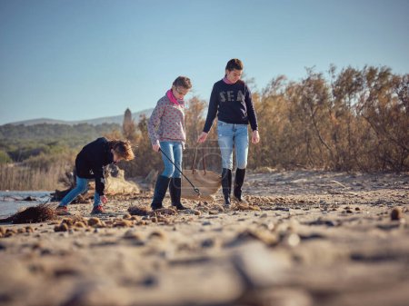 Foto de Cuerpo completo de niños en busca de basura y residuos con recogedor de basura en la costa arenosa del río en un día soleado sin nubes - Imagen libre de derechos