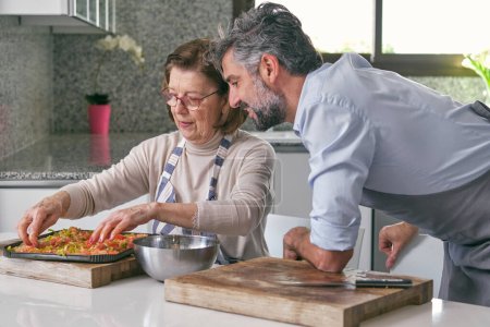 Photo for Side view of mature bearded Hispanic man with gray hair and elderly woman in aprons preparing delicious Italian food at home - Royalty Free Image
