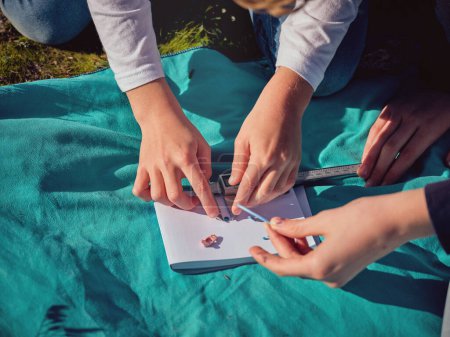 Photo for From above of crop anonymous preteen children sitting on grassy ground and measuring pieces on waste plastic material using digital caliper placed on sheet of notebook - Royalty Free Image