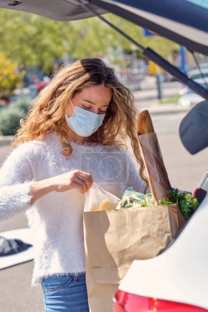 Téléchargez les photos : Jeune femme en vêtements décontractés et masque de protection plaçant des sacs d'épicerie dans le coffre de la voiture garée sur un parking hypermarché ensoleillé - en image libre de droit