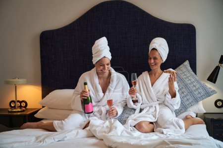 Photo for Cheerful female friends resting on bed in terry bathrobes drinking champagne and eating delicious croissants - Royalty Free Image