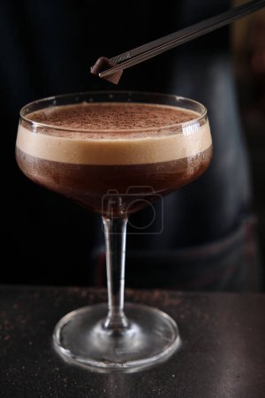 Foto de Barman irreconocible decorando cóctel de martini espresso espumoso con pieza de chocolate usando pinzas en el mostrador en el bar - Imagen libre de derechos