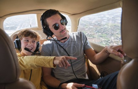 Photo for Excited little boy smiling in casual clothes and headset smiling and pointing away while sitting in aircraft cabin with young father during excursion trip - Royalty Free Image