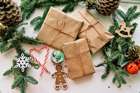 Photo for Top view of wrapped gift boxes with gingerbread man cookie and various Christmas decorations composed on while table with fir tree branches and cones - Royalty Free Image