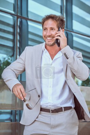 Téléchargez les photos : Entrepreneur masculin positif en costume élégant souriant tout en ayant une conversation téléphonique tenant des lunettes de vue dans une autre main regardant loin debout à l'intérieur - en image libre de droit