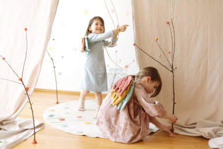 Foto de Adorables niñas con coloridos trajes de alas de ángel jugando con palos en el suelo de madera por la cortina blanca sobre la estera de algodón suave en la sala de luz - Imagen libre de derechos