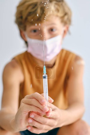 Foto de Niño con mascarilla en la cara mirando a la cámara mientras rocía la vacuna del inyector durante la pandemia de COVID 19 - Imagen libre de derechos