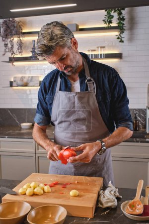 Téléchargez les photos : Homme barbu concentré dans le tablier debout près du comptoir de la cuisine et peler la tomate sur la planche à découper - en image libre de droit