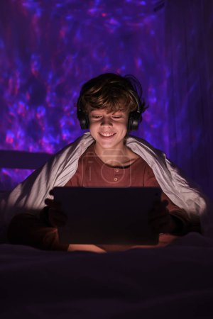 Téléchargez les photos : Contenu enfant en pyjama et écouteurs regarder vidéo intéressante sur tablette tout en étant assis sur le lit dans la chambre sombre la nuit - en image libre de droit