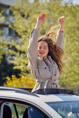 Photo for Joyful attractive female in warm coat standing with eyes closed in car sunroof and raising arms in excitement on sunny spring day - Royalty Free Image