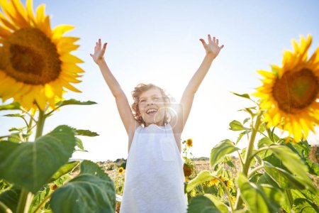 Foto de Bajo ángulo de niño feliz en camisa sin mangas con el pelo rizado levantando los brazos mientras está de pie en la floreciente pradera de girasol y disfrutando del cálido clima soleado de verano - Imagen libre de derechos