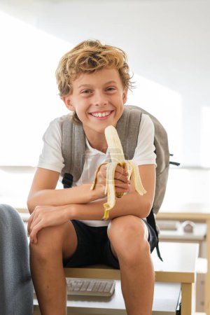 Fröhliches Schulkind mit Rucksack sitzt auf dem Tisch im Klassenzimmer und isst in der Pause Banane, während er in die Kamera schaut