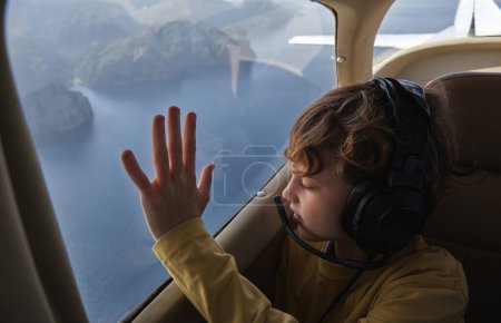 Foto de Lindo chico en auriculares volando en biplano y mirando por la ventana con interés mientras vuela sobre el mar azul y la costa rocosa - Imagen libre de derechos