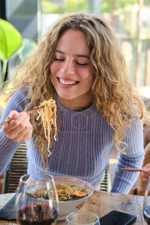 Téléchargez les photos : Une jeune femme positive assise à table et mangeant des ramen épicés avec des baguettes dans un restaurant moderne pendant le week-end - en image libre de droit