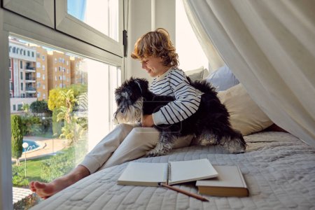Photo for Glad schoolkid with crossed legs interacting with Miniature Schnauzer on bed with textbook and notepad at home - Royalty Free Image